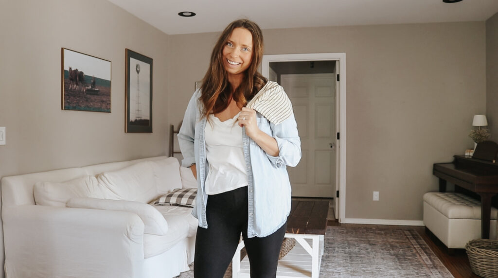 Christian homemaker stands in living room wearing a white shirt and a light blue overshirt with black pants